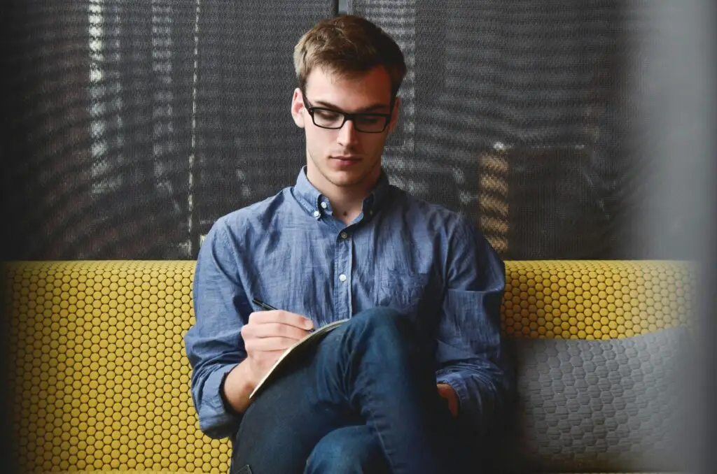 A young man wearing glasses, seated on a yellow patterned sofa, focusing intently while writing in a notebook—representing concentration, pattern recall, and cognitive memory skills.
