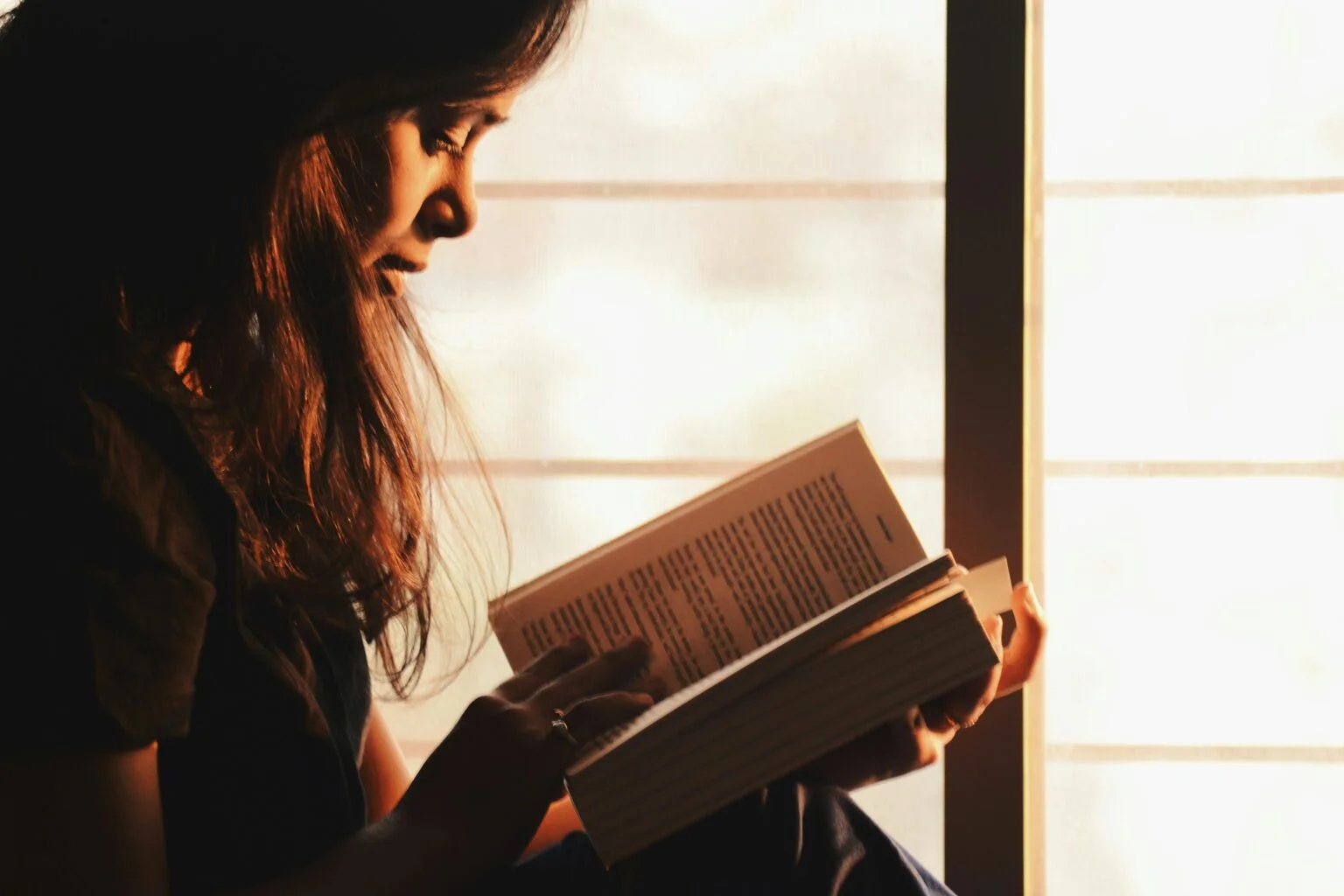 Woman reading a book in warm sunlight near a window, focused and immersed in text, representing the cognitive and memory benefits of reading.