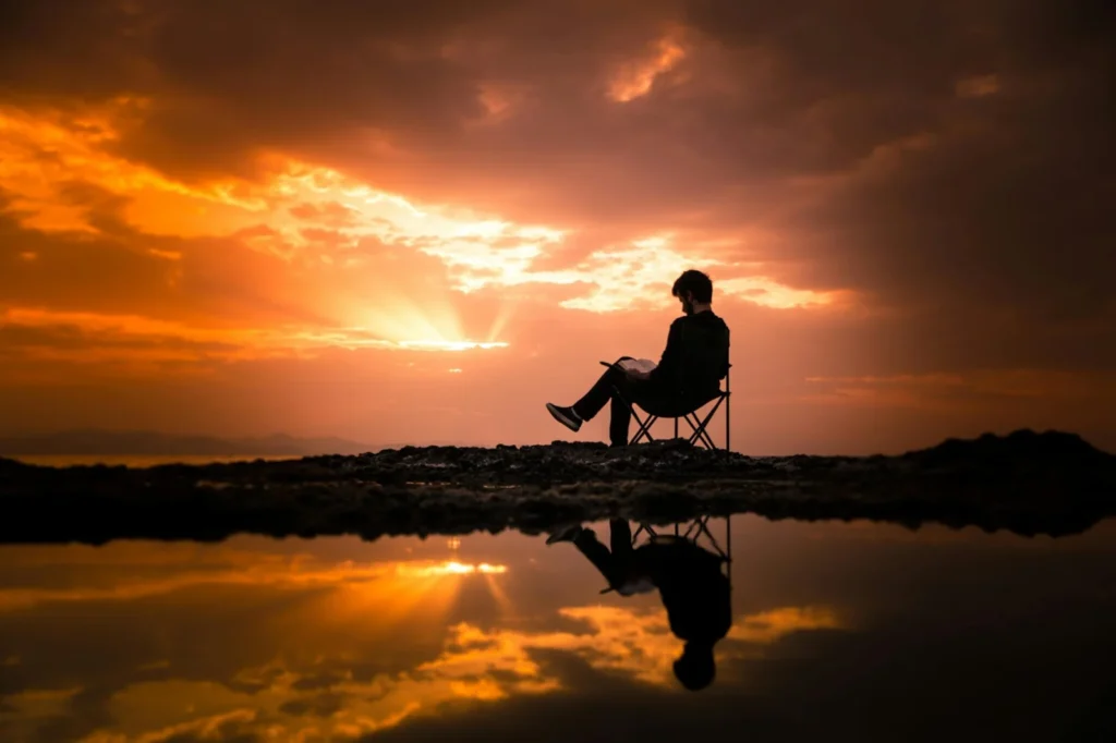 “Person reading a book outdoors during a dramatic sunset, seated in a chair by reflective water, symbolizing mindful reading and cognitive relaxation.”